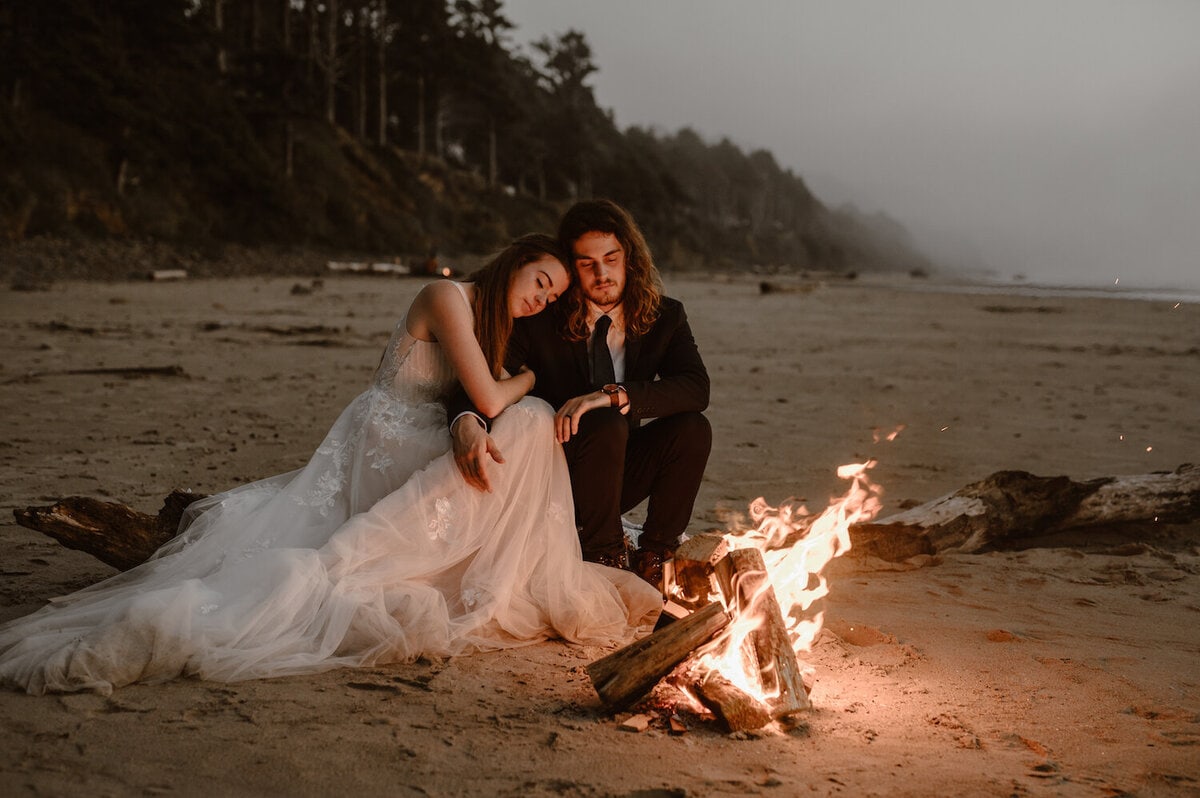a man and woman sitting next to a fire on a beach