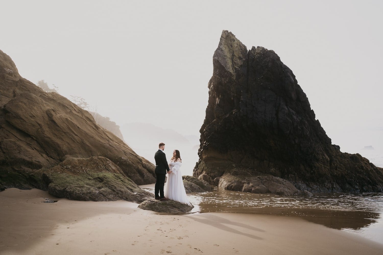 A landscape photo showing a bride and groom standing together between the rocks at Hug Point near Cannon Beach.