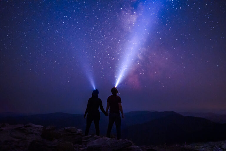 A couple is silhouetted by the milkyway. Their headlamps shoot beams of white light into the sky